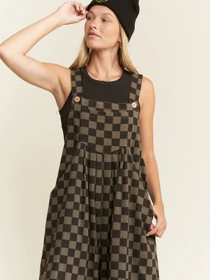 Model wearing wide leg checkerboard overalls with adjustable straps, front pocket, and black sleeveless top, accessorized with a black beanie.