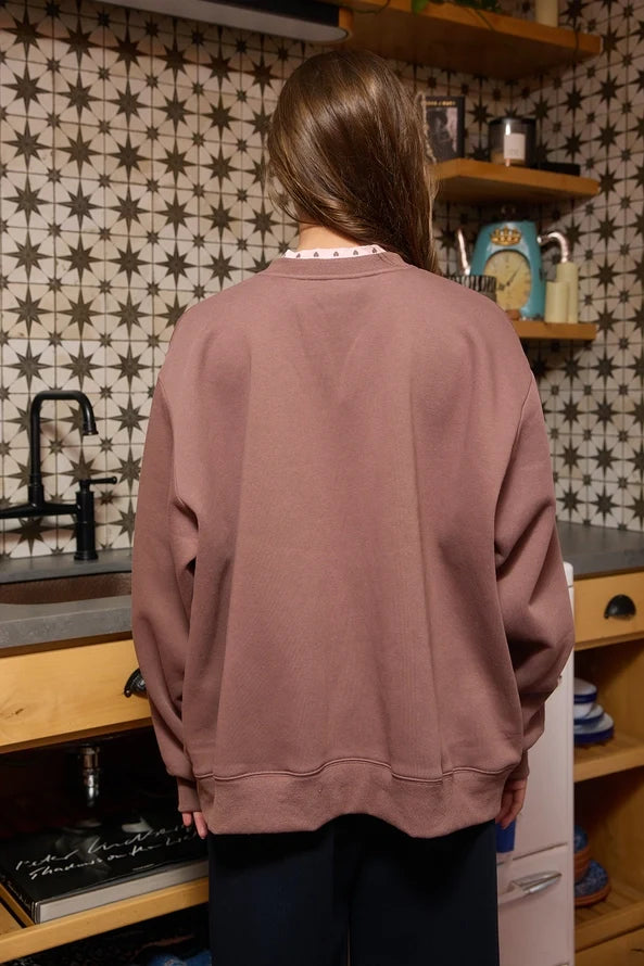 Back view of the brown Jesus sweatshirt showing relaxed fit and plain design, worn by a woman in a cozy kitchen with star-patterned wallpaper.