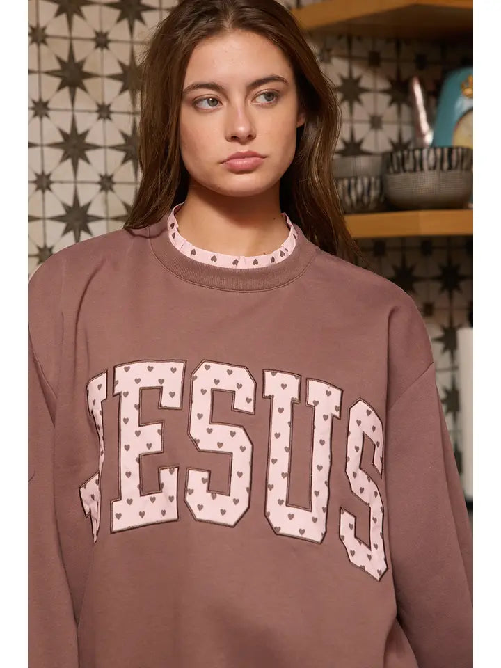 Close-up of the brown Jesus sweatshirt featuring the 'JESUS' heart appliqué with pink hearts, worn by a woman with long hair in a patterned kitchen.