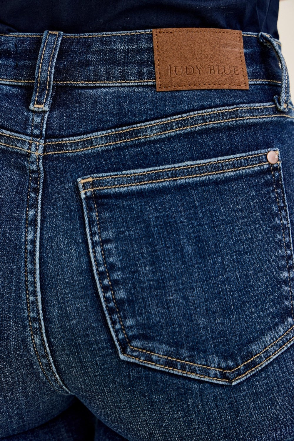 Close-up of dark blue denim fabric with contrast stitching, leather patch with 'Judy Blue' branding on high-rise petite flare jeans