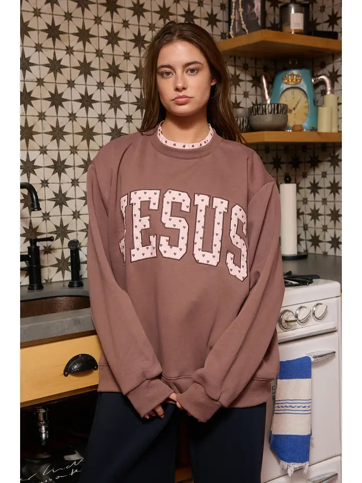 Front view of the oversized brown Jesus sweatshirt with 'JESUS' heart appliqué, styled with casual dark pants, in a kitchen setting.