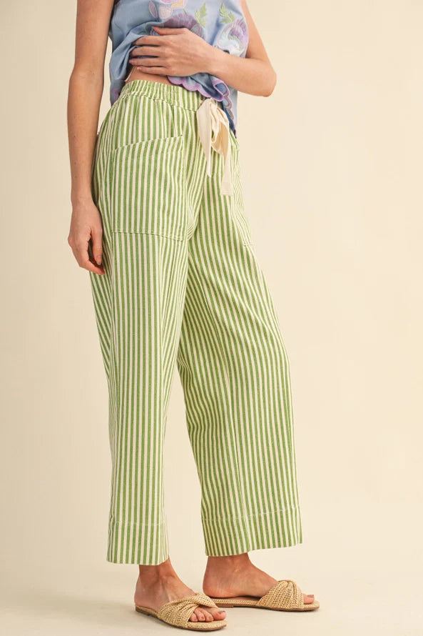 A model wearing light green and white vertically striped cropped pants with a cream drawstring, styled with a blue patterned top and beige slide sandals against a solid beige background.