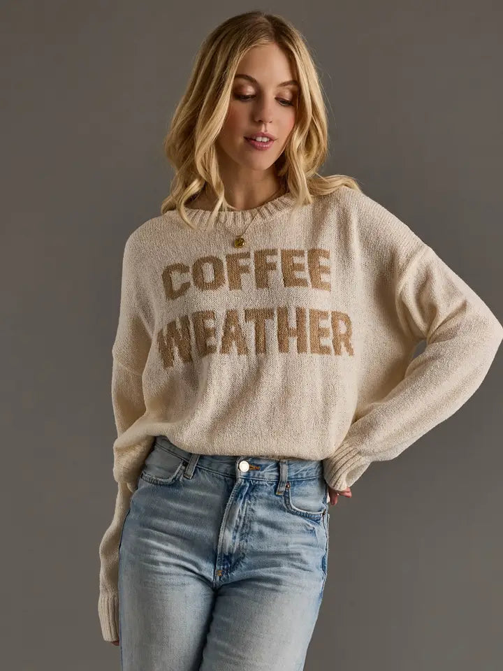Woman wearing an ivory Coffee Weather graphic knit sweater with taupe brown lettering, crew neckline, long sleeves, and relaxed fit styled with light wash jeans.
